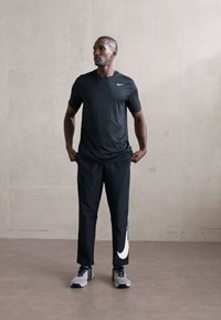 Atletisk mand i sort Nike skjorte og bukser med stor hvid swoosh, stående indendørs mod en simpel lys væg og ser til siden.