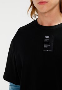Schwarzes Baumwoll-T-Shirt mit Rundhalsausschnitt und kurzen Ärmeln. Enthält ein Markenlabel mit weißem Text auf der Vorderseite.