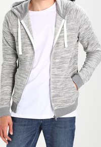 Haut à zip gris avec une finition texturée, cordons de serrage blancs et un bord contrastant gris clair. Porté sur un t-shirt blanc.