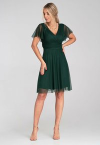 Dunkelgrünes kurzes Kleid mit durchsichtigen Flatterärmeln, einem V-Ausschnitt und einem taillierten Oberteil. Der Rock weitet sich sanft und hat eine fließende, leichte Textur.