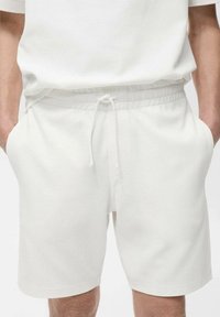 Shorts en coton blanc avec une taille élastique et un cordon de serrage, dotés de poches latérales et d'une texture lisse. Design simple et fonctionnel.