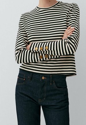 Personne portant une chemise à manches longues rayée noir et blanc avec des boutons dorés aux poignets et un jean taille haute bleu foncé, bras croisés.