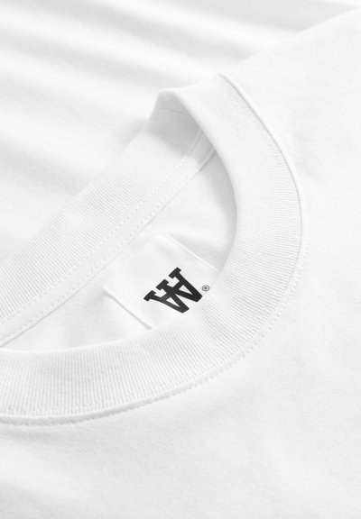 Κοντινό πλάνο στον λαιμό ενός λευκού T-shirt με ραβδωτό γιακά τύπου crew και ορατή λευκή ετικέτα που φέρει μαύρο στυλιζαρισμένο λογότυπο "AA".