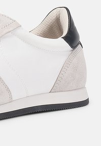 Hvit sneaker med grå suedeforsterkninger, glatt tekstiloverdel, svart hæltab, gummisåle og synlig søm. Klassisk atletisk design.
