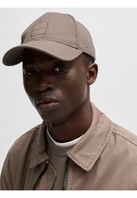 Cappello da baseball beige con patch quadrato, abbinato a un giubbotto beige sopra un maglione chiaro. Tessuto morbido, design strutturato, palette di colori neutri.