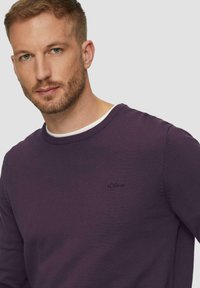 Violeta adīta džemperis ar apaļu kakla izgriezumu un ribotām piedurknēm, ar mazu melnu logotipu uz krūšu. Gluda tekstūra.