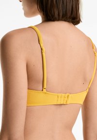 Reggiseno giallo con texture, dotato di spalline regolabili e chiusura a gancio, caratterizzato da un pannello posteriore liscio e dettagli hardware minimi.