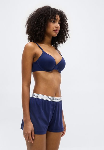 Γυναίκα με σγουρά μαλλιά που φοράει μπλε σουτιέν σε ναυτικό μπλε και αντίστοιχα σορτς Polo Ralph Lauren, στέκεται μπροστά σε απλό φωτεινό φόντο.