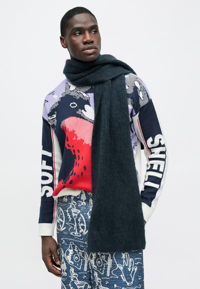 Σκούρα μπλε μάλλινο κασκόλ τυλιγμένο γύρω από τον λαιμό, σε συνδυασμό με πολύχρωμο πουλόβερ με γραφικό σχέδιο και παντελόνια με σχέδιο.