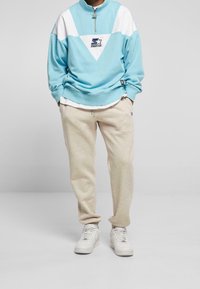 Ljusblå zip-up sweatshirt med vita accenter, med en triangulär design och logotyp. Kombinerad med beige sweatpants och vita sneakers.