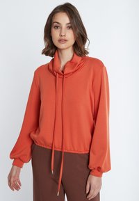 Pullover mit Kapuze und langen Ärmeln aus orangerem Stoff mit Kordelzugsaum. Glatte Textur; ausgestellt mit braunen Hosen.