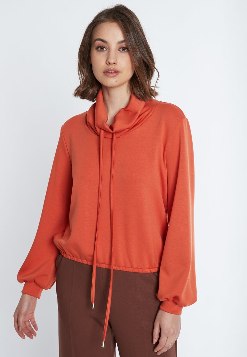 Pullover mit Kapuze und langen Ärmeln aus orangerem Stoff mit Kordelzugsaum. Glatte Textur; ausgestellt mit braunen Hosen.