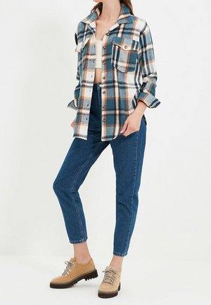 Femme portant une chemise en flanelle à carreaux bleue, blanche et orange sur un crop top blanc, un jean bleu taille haute et des chaussures de randonnée beige.