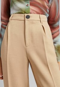 Beige, hoch taillierte Hose mit Falten, ausgestattet mit einer vorderen Tasche und einem schwarzen Knopfverschluss, aus einem glatten Stoff gefertigt.