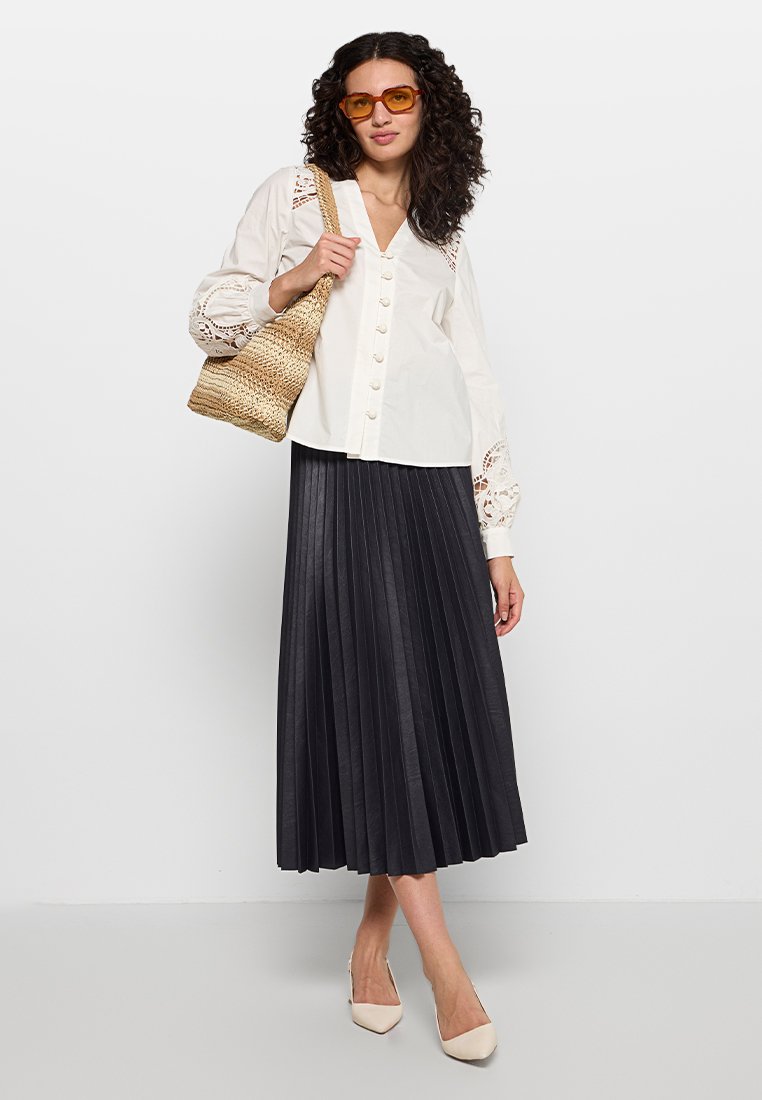 Weißes Hemd mit Spitzenakzenten, schwarzer plissierter Midi-Rock, beige spitze Pumps und geflochtene Strohtasche. Orangefarbene Sonnenbrille.