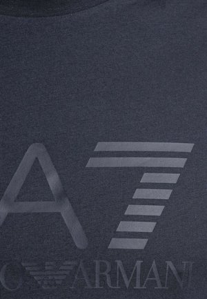 Dunkler Stoff mit großem, stilisiertem "A7" und schwachem "Armani"-Logo mit einem Adleremblem unter den Buchstaben.