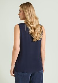 Mujer con cabello largo y ondulado rubio, vestida con una camiseta sin mangas azul marino y pantalones a juego, de perfil contra un fondo claro y liso.