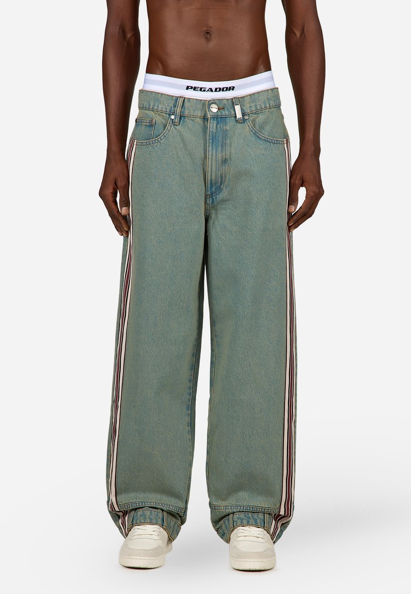 Svijetloplave široke traperice izrađene od denima, s dvije vertikalne pruge u crvenoj i bijeloj boji s obje strane, s bijelim PEGA-DOR pojasom.