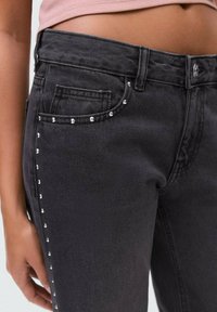 Črne jeans hlače v oprijetem kroju, s kamenčkastim teksturo in srebrnimi zakovicami ob stranskih šivih in robovih žepov za dodaten detajl.