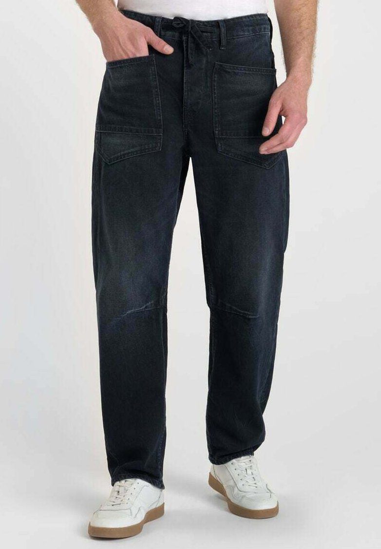 Homme portant un pantalon en jean noir avec de grandes poches avant et des baskets blanches, debout sur un fond clair uni.
