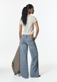 Kratek rokav svetlo sivi top in široke modre jeans hlače z rjavim obližem. Model drži olivno zeleno jakno in stoji ob enotni ozadju.