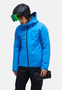Helderblauw waterdicht ski-jas met een capuchon, zwarte rits en logo. Gecombineerd met een zwarte broek en het vasthouden van zwarte wanten.