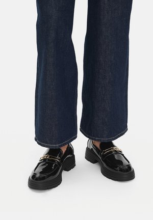 Mocassini in pelle verniciata nera con dettagli in oro e suole spesse in gomma nera, indossati con pantaloni a gamba larga in denim scuro.