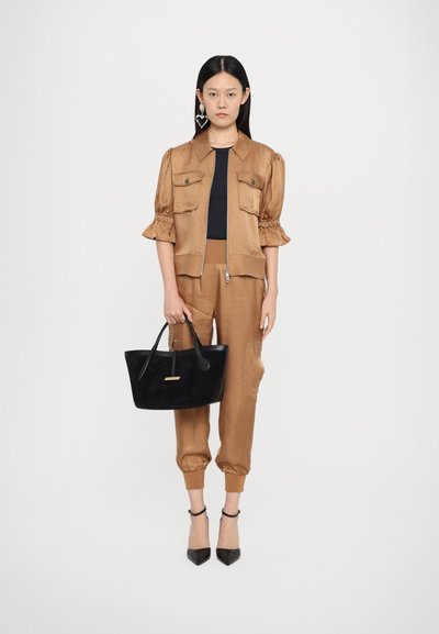 Braune satinierte Kurzjacke mit Puffärmeln und vorderen Taschen, passende braune Hose, schwarze Absatzschuhe und eine schwarze Handtasche mit goldenen Akzenten.