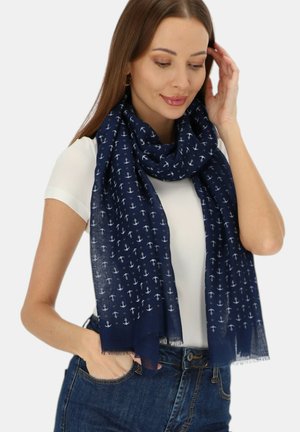 Kobieta ubrana w granatowy szalik z białymi wzorami kotwicy, białą bluzkę z krótkim rękawem i niebieskie dżinsy, dotykająca włosów jedną ręką.