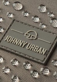 Tessuto grigio con una superficie texturizzata, caratterizzato da gocce lucide e una toppa rettangolare con il logo "JOHNNY URBAN" in rilievo beige.