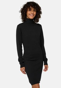 Lächelnde Frau mit lockigem Haar, die ein schwarzes, langärmliges Turtleneck-Strickkleid vor einem schlichten hellen Hintergrund trägt.