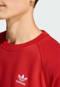 Crvena dukserica od teksturiranog materijala, s okruglim izrezom i bijelim Adidas logom. Vidljive šavove na ramenu.