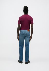 Tričko s krátkym rukávom bordovej farby a svetlomodré džínsové nohavice. Na zadnej strane džínsov je kožená nášivka s logom. Viditeľné čierne topánky. Neutrálne pozadie.