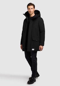 Melnā pūkaina jaka ar kapuci, priekšpusē ar rāvējslēdzēju, kabatas ar atloku un logotipa ielāpu. Valkāta kopā ar tumšām džinsām un melniem zābakiem.