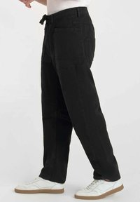 Zwarte, loszittende broek met een tailleband met trekkoord. Heeft grote zijzakken en een gladde, duurzame stof. Gepaard met witte sneakers.