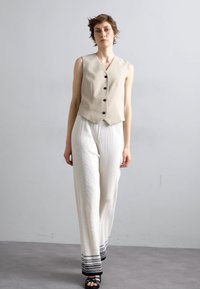 Beige mouwloze vest met knopen, gecombineerd met witte geplooide broek met een zwarte patroonrand. Het model draagt zwarte sandalen.