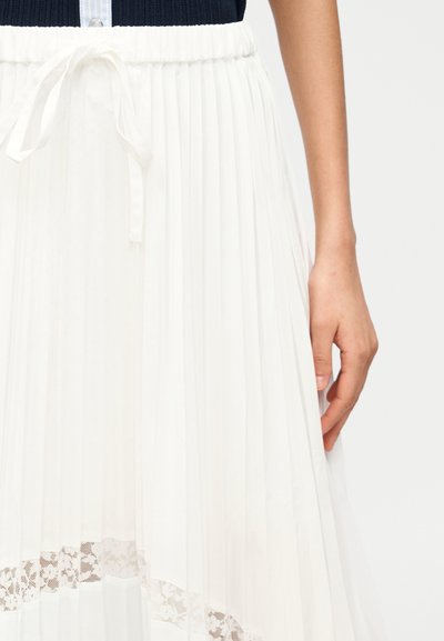 Χέρι δίπλα σε λευκή πλισέ φούστα με λεπτομέρεια από δαντέλα στο τελείωμα και δεμένη μέση, φορεμένη με μπλε σκούρο τοπ.