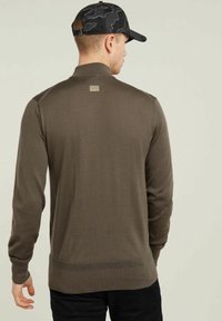 Maglione marrone a maglia con maniche lunghe, colletto alto, texture a coste e dettaglio del logo sul retro. Indossato con un berretto nero a fantasia.