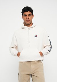 Felpa color crema con tasca frontale, cappuccio regolabile e un patch con logo. Presenta una tipografia sulla manica in blu scuro.