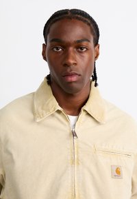 Veste zippée en velours côtelé beige avec un col chemise, une poche avant et un patch logo. Texture lisse et design moderne.