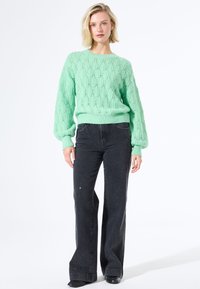 Vrouw met kort blond haar, draag een mintgroene gebreide trui en hoge zwarte wijde broek tegen een witte achtergrond.