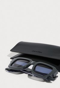 Šedé obdélníkové sluneční brýle s tmavými čočkami, výrazným rámečkem a průhlednými detaily, položené na černém obalu Calvin Klein.