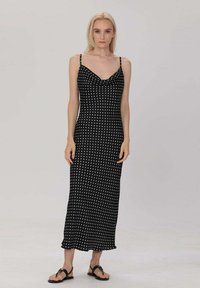 Robe maxi noire sans manches avec des pois blancs, design à col drapé, coupe slim et ourlet volanté. Portée avec des sandales plates.
