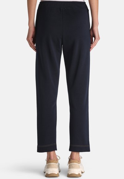 Persona vistiendo pantalones negros de pierna recta y zapatillas beige, de pie de espaldas contra un fondo claro y liso.