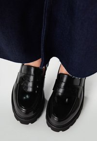 Zwarte gepoetste leren instappers hebben een dikke zool en een band over de voorkant met uitsparingen. Geweven stiksels langs de randen.