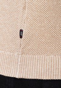 Ourlet de pull en maille beige avec motif texturé et petite étiquette en tissu noir portant le nom "JEFF" au-dessus de la bande côtelée, associé à un pantalon noir.