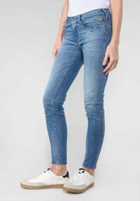 Mujer con jeans ajustados azul claro y zapatillas blancas con detalles en beige y negro, de pie contra un fondo claro y liso.