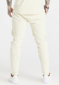 Kremowe, obcisłe joggery wykonane z miękkiego materiału, z tapered design, elastycznym pasem oraz mankietami na kostkach, w połączeniu z białymi sneakersami.