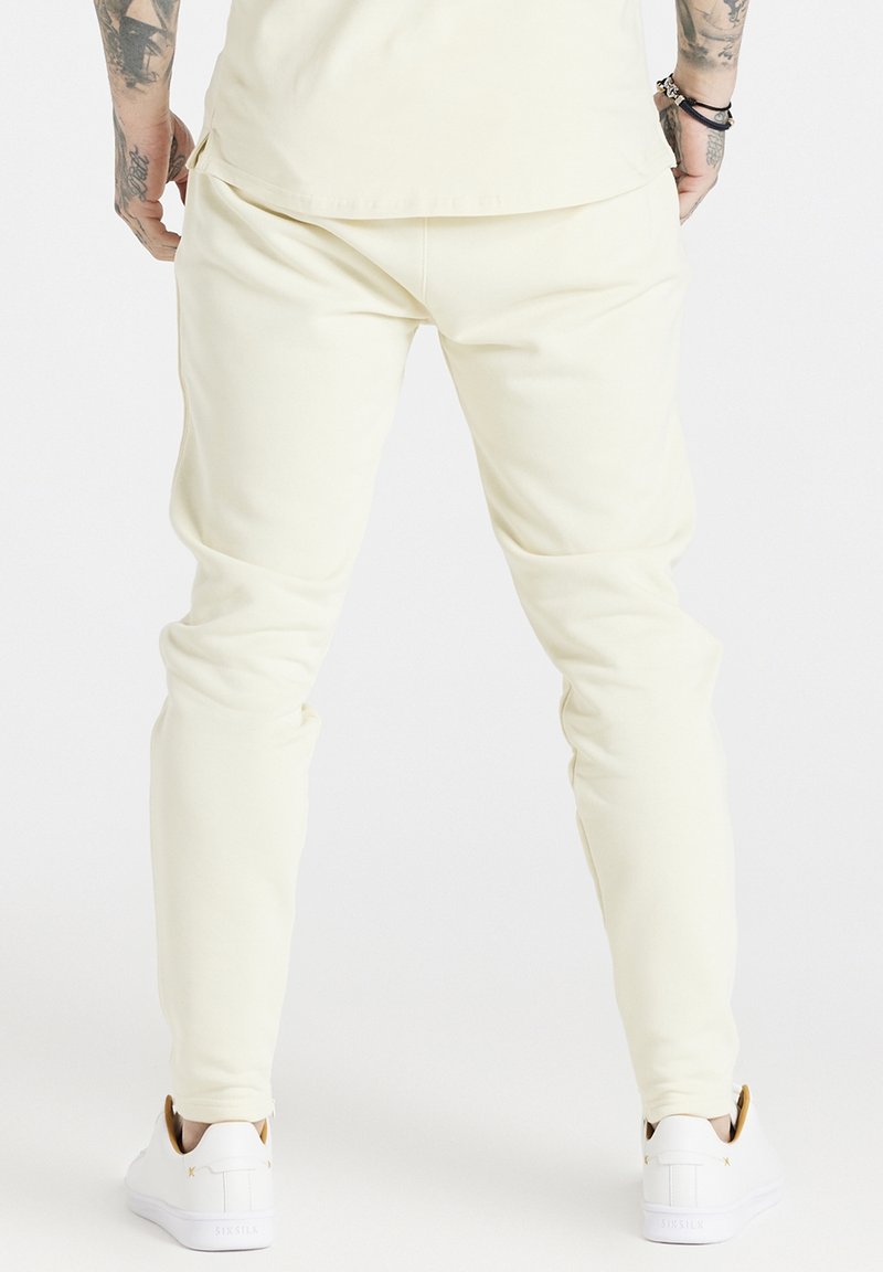 Kremowe, obcisłe joggery wykonane z miękkiego materiału, z tapered design, elastycznym pasem oraz mankietami na kostkach, w połączeniu z białymi sneakersami.