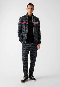 Trainingsanzug in Marineblau mit Logomuster, bestehend aus einer Zip-up-Jacke mit roten und weißen Streifen und passenden Hosen. Schwarze Sneakers vervollständigen den Look.